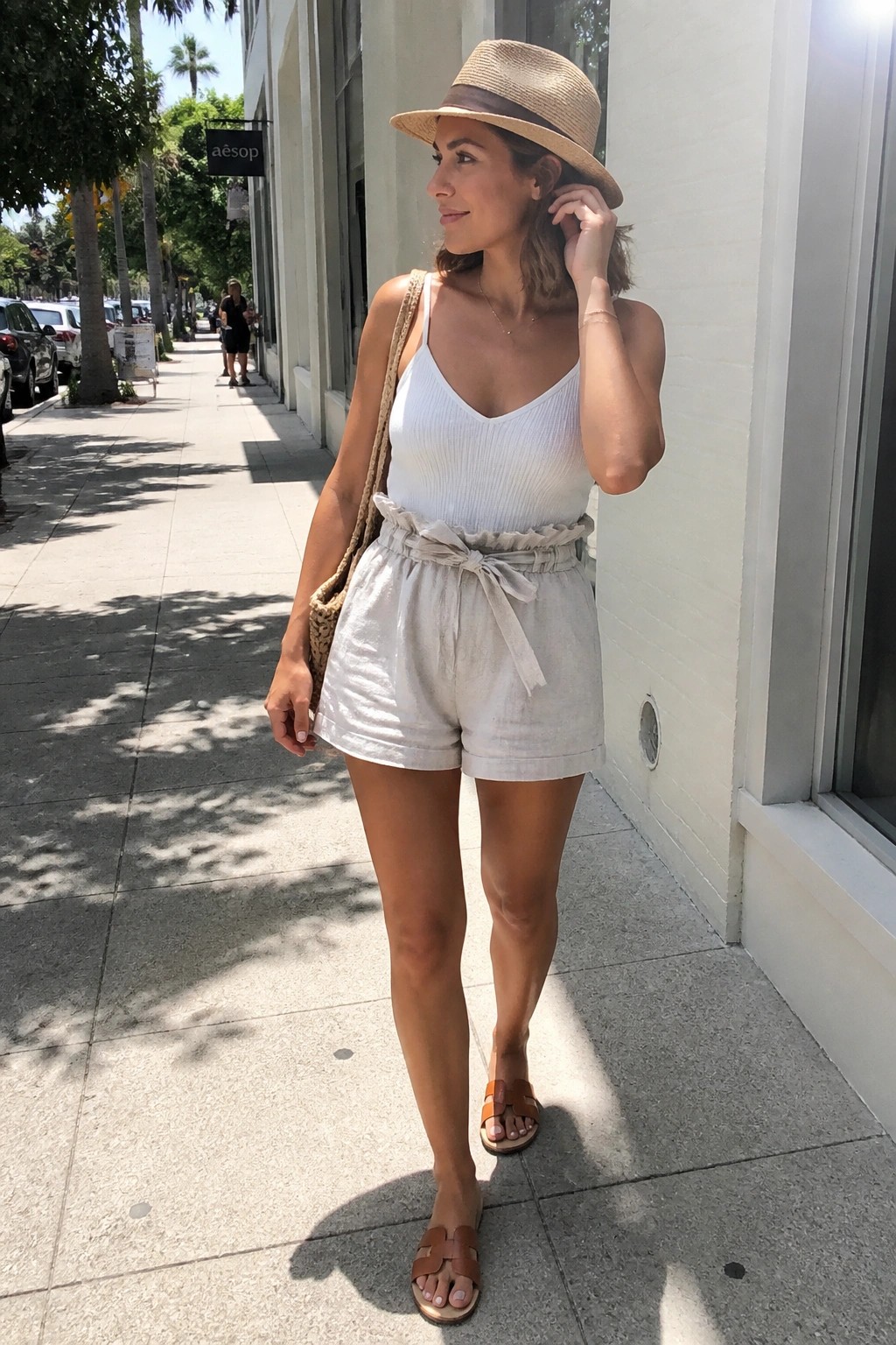 Woman in white ribbed camisole top, light beige linen shorts with tie waist, straw fedora hat, brown slide sandals, and woven shoulder bag standing on a sidewalk.