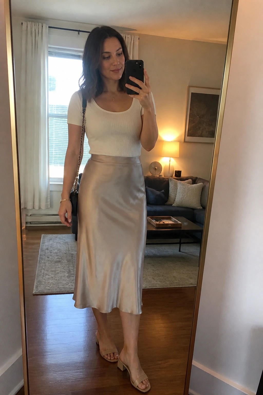 A woman wearing a cream ribbed short sleeve top with a beige satin midi skirt, beige block heel sandals, and a black handbag with gold chain strap, standing in a living room taking a mirror selfie.