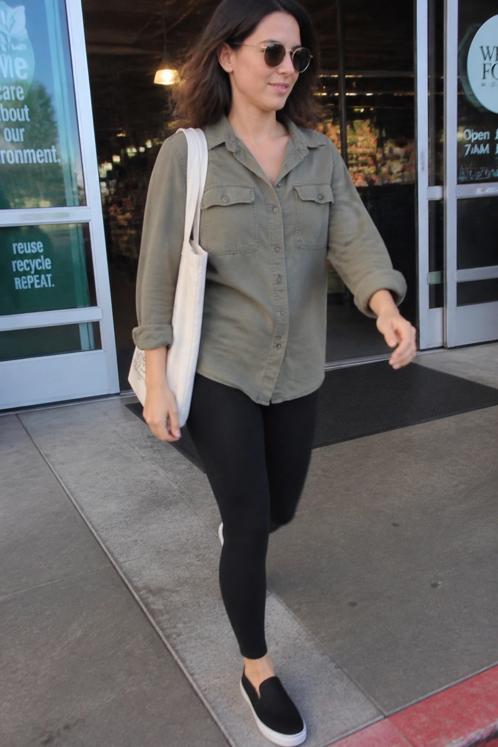 Woman wearing an olive green button-up utility shirt over black leggings with black slip-on sneakers and a white tote bag slung over one shoulder.