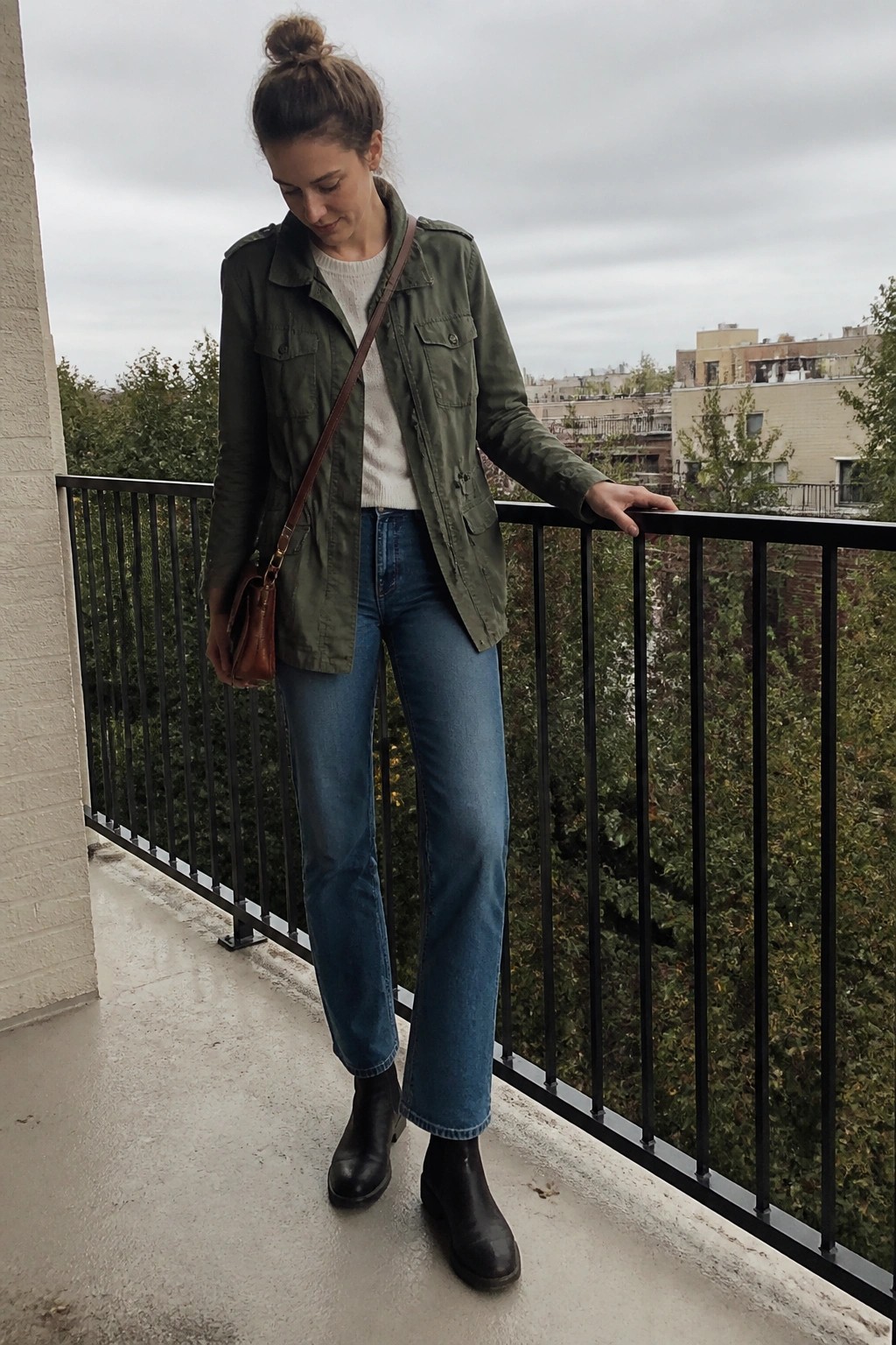 Woman standing on a balcony wearing an olive green utility jacket over a cream sweater with blue straight-leg jeans, black ankle boots, and a brown leather crossbody bag.