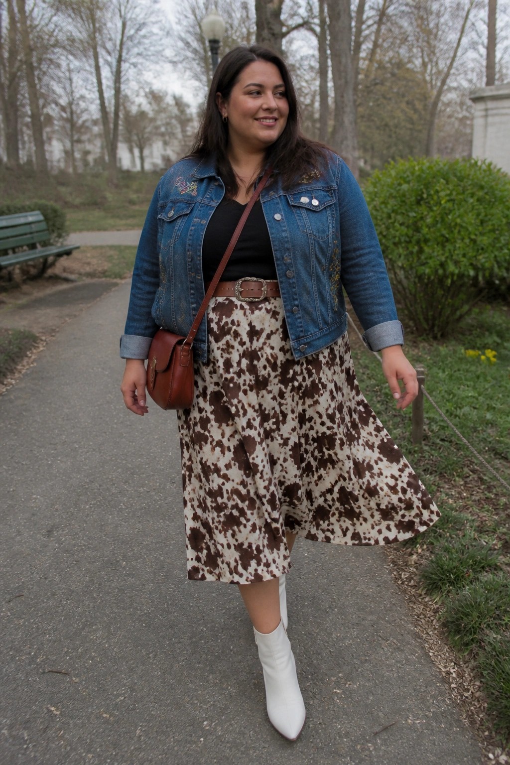 Woman in blue denim jacket and cow-print skirt walks in park wearing white boots.