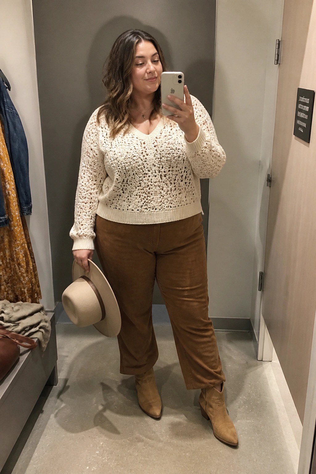 Woman in cream open-knit sweater and brown corduroy pants taking mirror selfie in fitting room