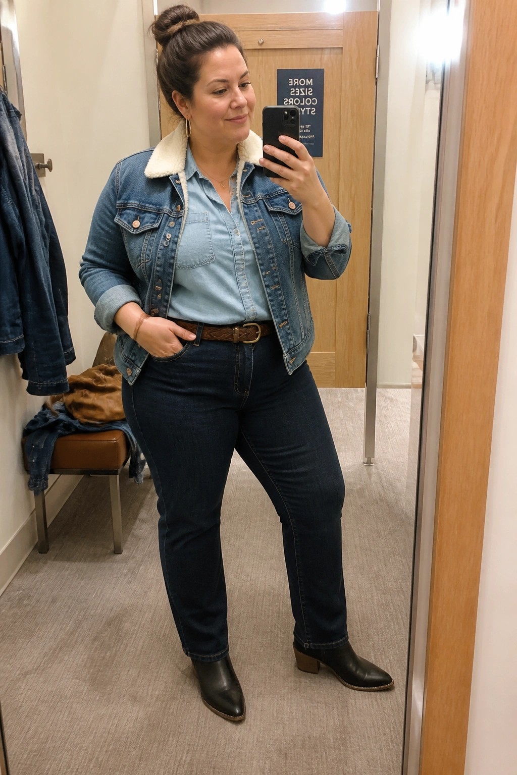 Woman in denim jacket with shearling collar, light blue shirt, and dark jeans taking mirror selfie