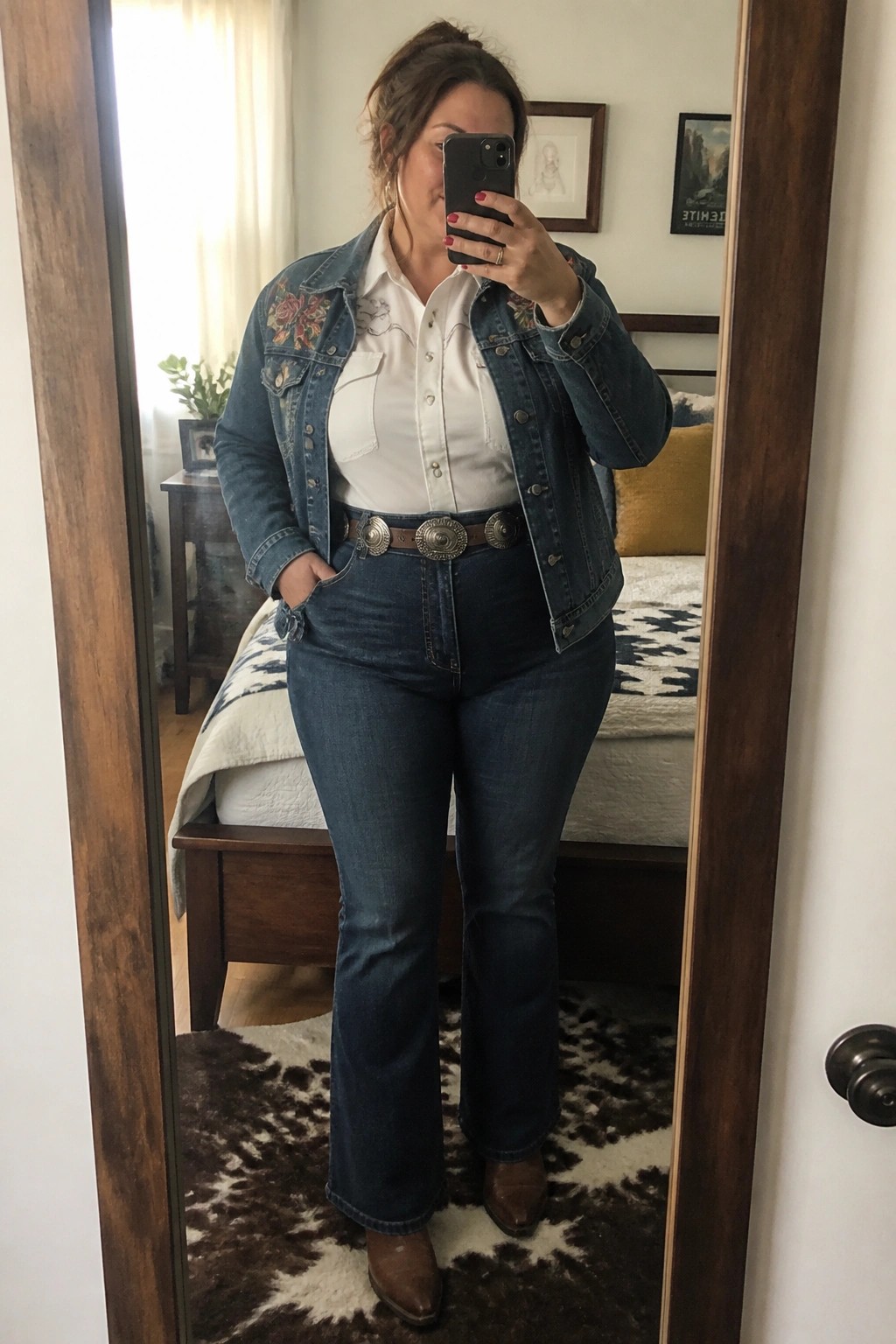 Plus size woman wearing a blue denim jacket with floral embroidery on the shoulders over a white button down shirt, paired with dark blue flared jeans, a brown leather belt featuring large silver conchos, and brown pointed toe cowboy boots.