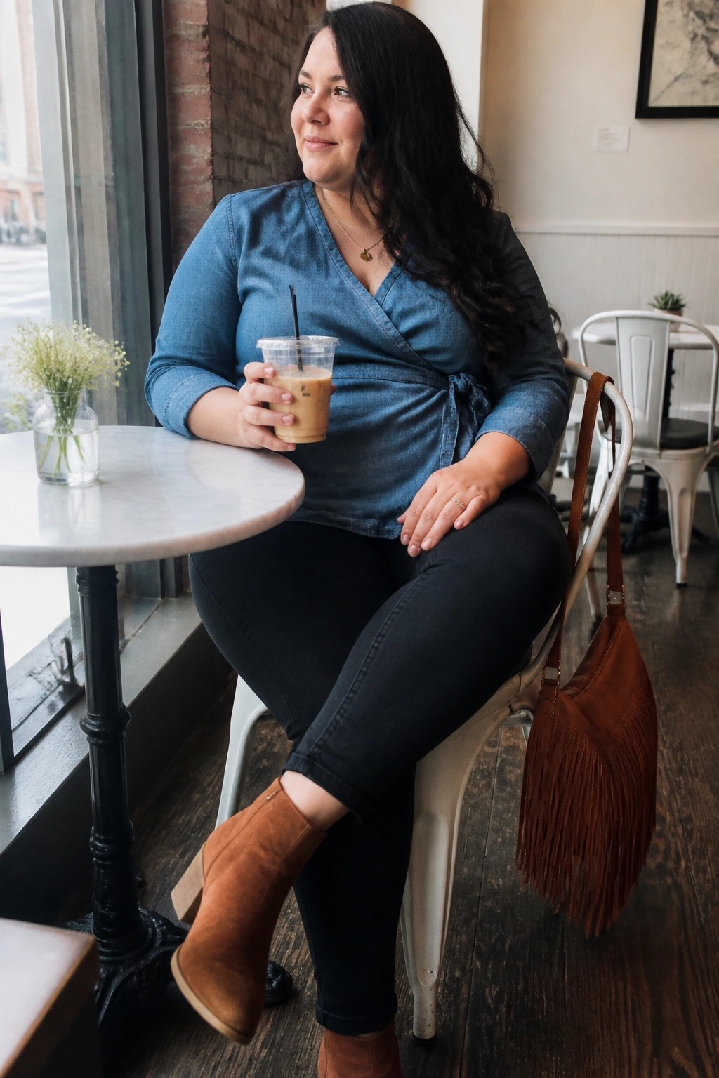 Woman in blue denim wrap top holding iced coffee in cafe chair