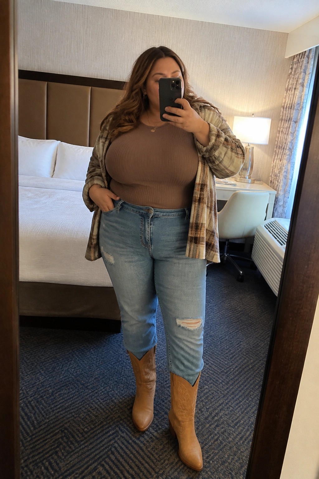 Woman in brown top, plaid flannel, ripped jeans, and cowboy boots in hotel room