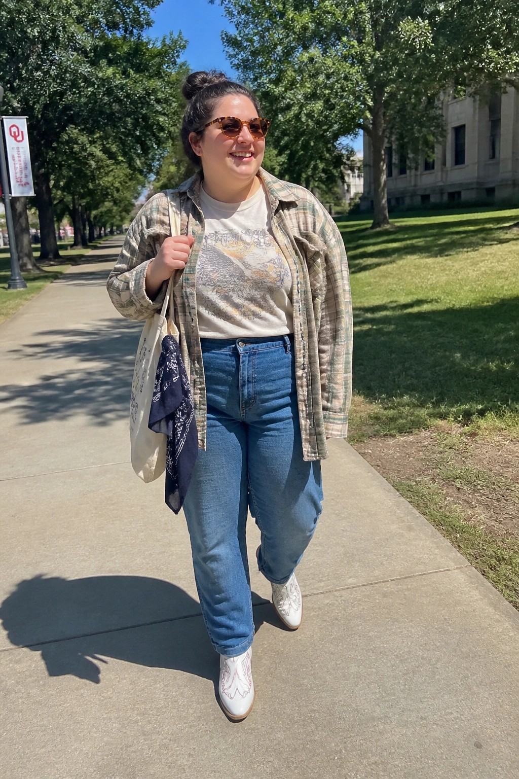 Plus size woman wearing an open plaid flannel shirt over a mountain graphic tee paired with blue jeans, white embroidered cowboy boots, and a canvas tote bag with a navy bandana.