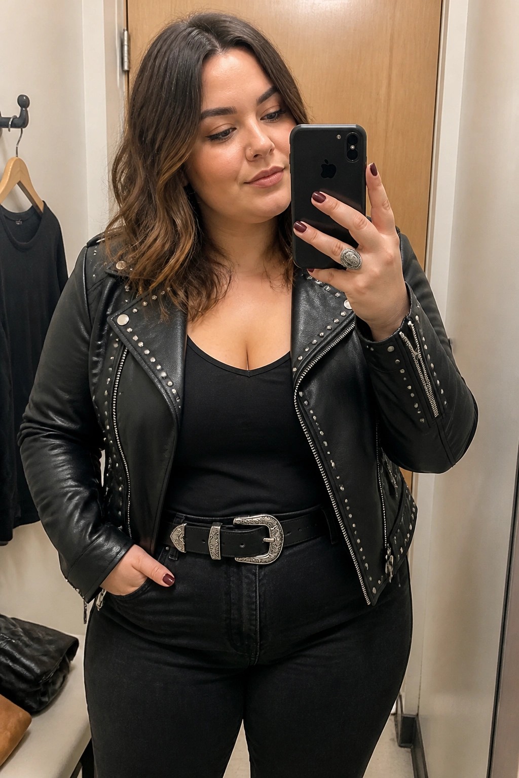 Woman in black studded leather jacket taking mirror selfie with iPhone in fitting room