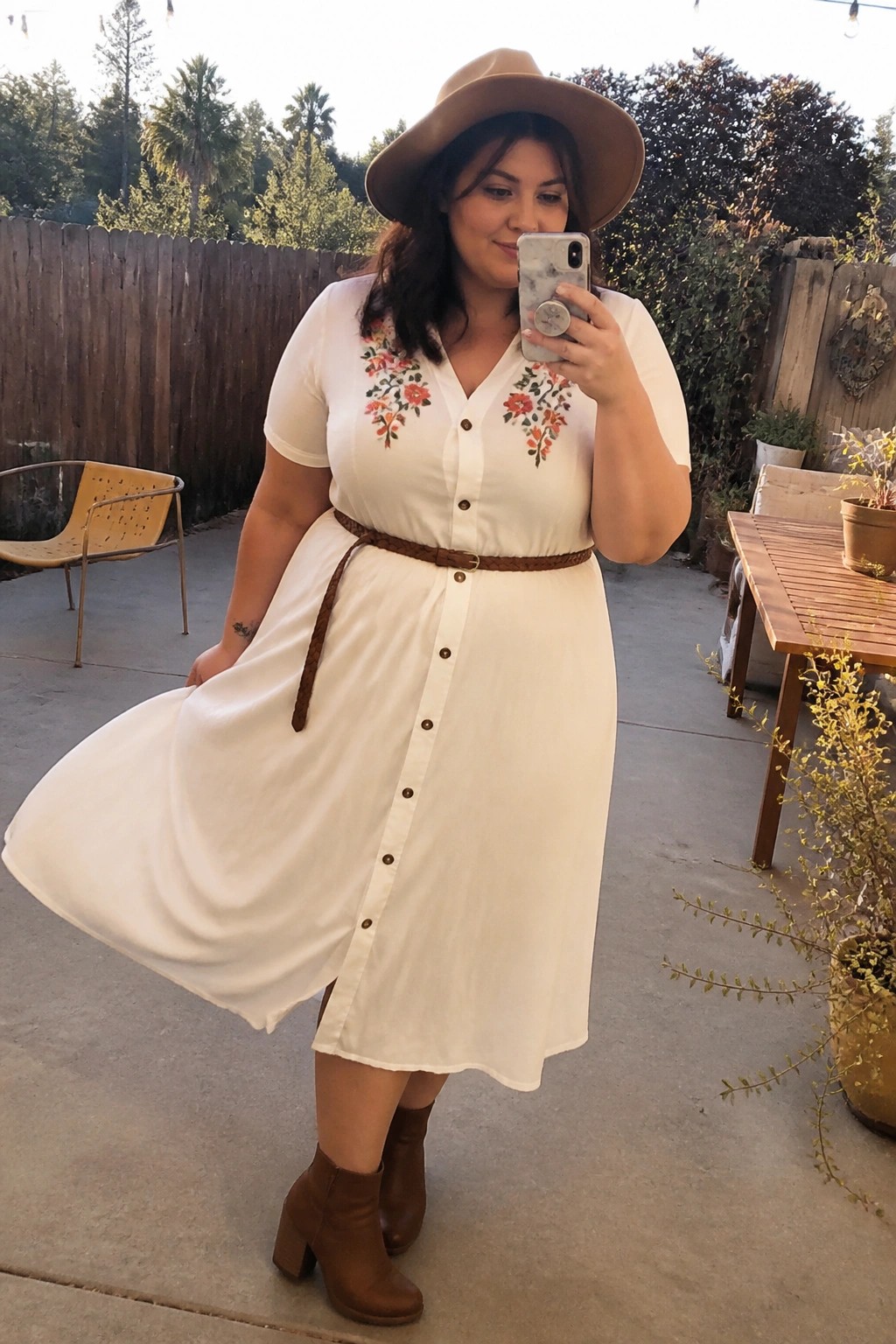 Woman in white floral-embroidered dress, brown hat and boots taking selfie outdoors