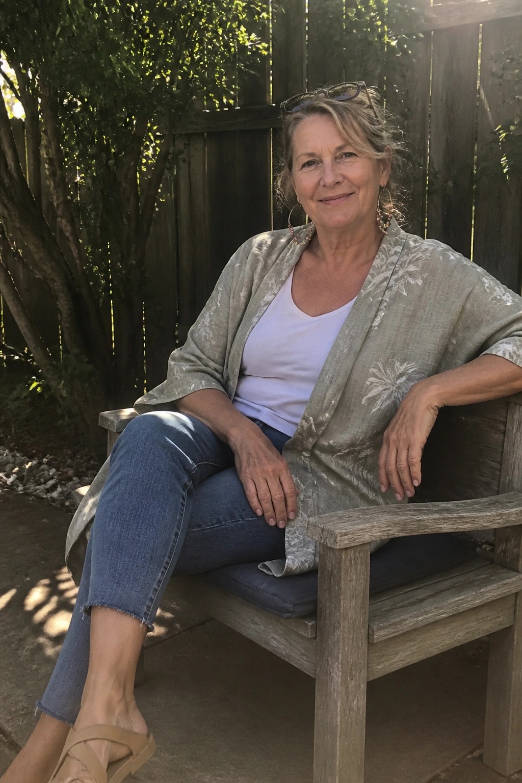 Woman over 50 seated outdoors wearing a sage green patterned linen jacket open over a white cotton tank top, blue cuffed jeans, and tan flat sandals.