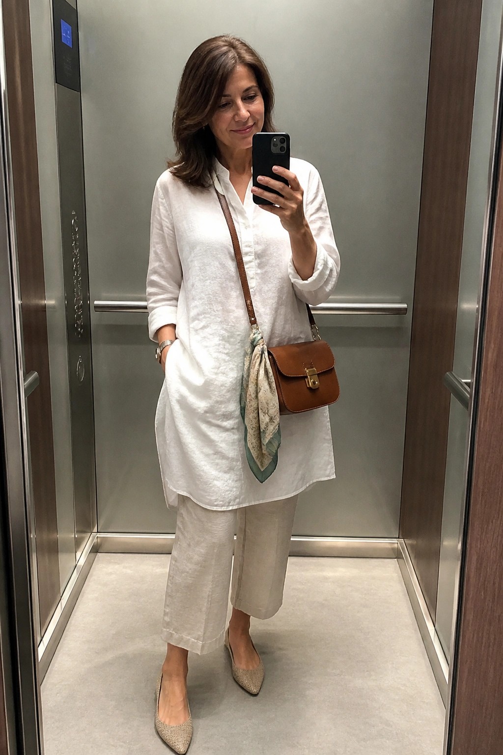 A woman in an elevator taking a mirror selfie while wearing a white linen button-down shirt, light beige linen cropped pants, sparkly pointed flats, and a brown leather crossbody bag with a patterned scarf tied on.