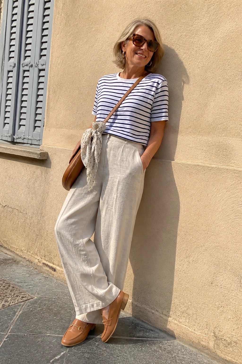 Smiling woman in striped tee and white wide-leg pants leaning against beige wall with brown bag.