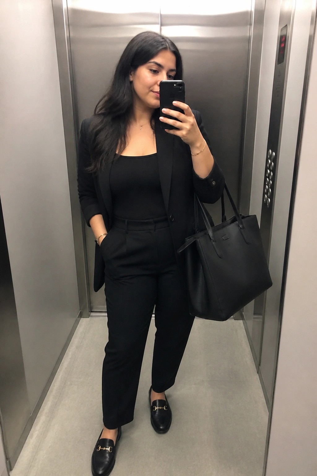 Woman wearing a black blazer over a black square neck top with black straight leg trousers and black loafers holding a black tote bag in an elevator.