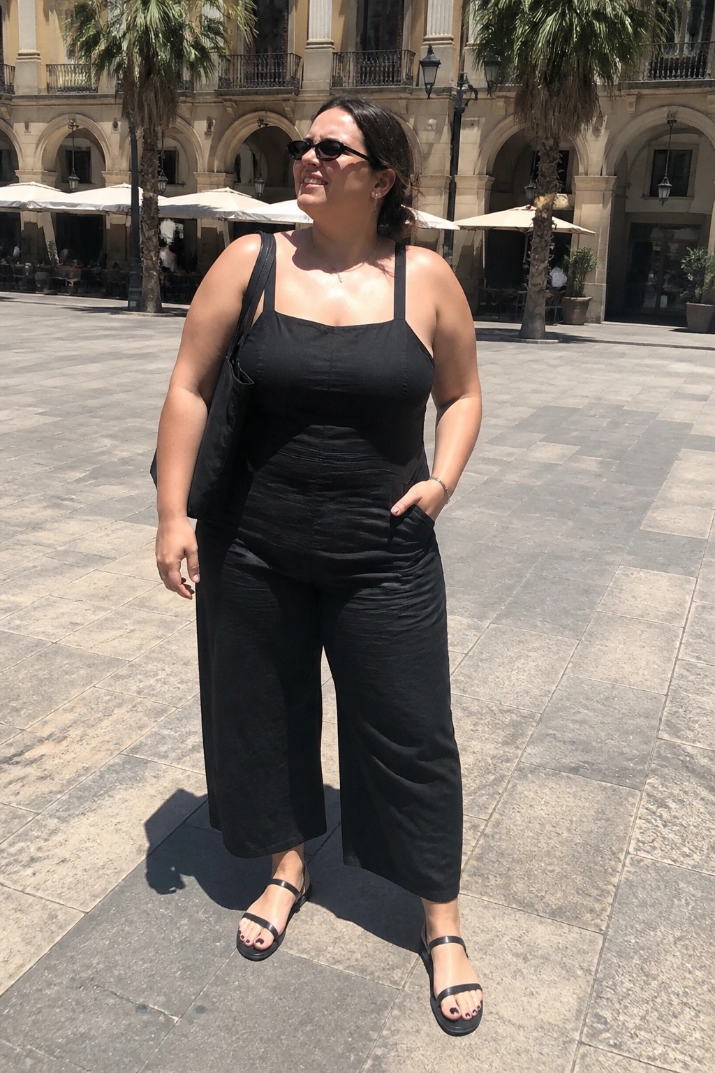 Woman in black top and wide pants stands in sunny plaza with palms and arches