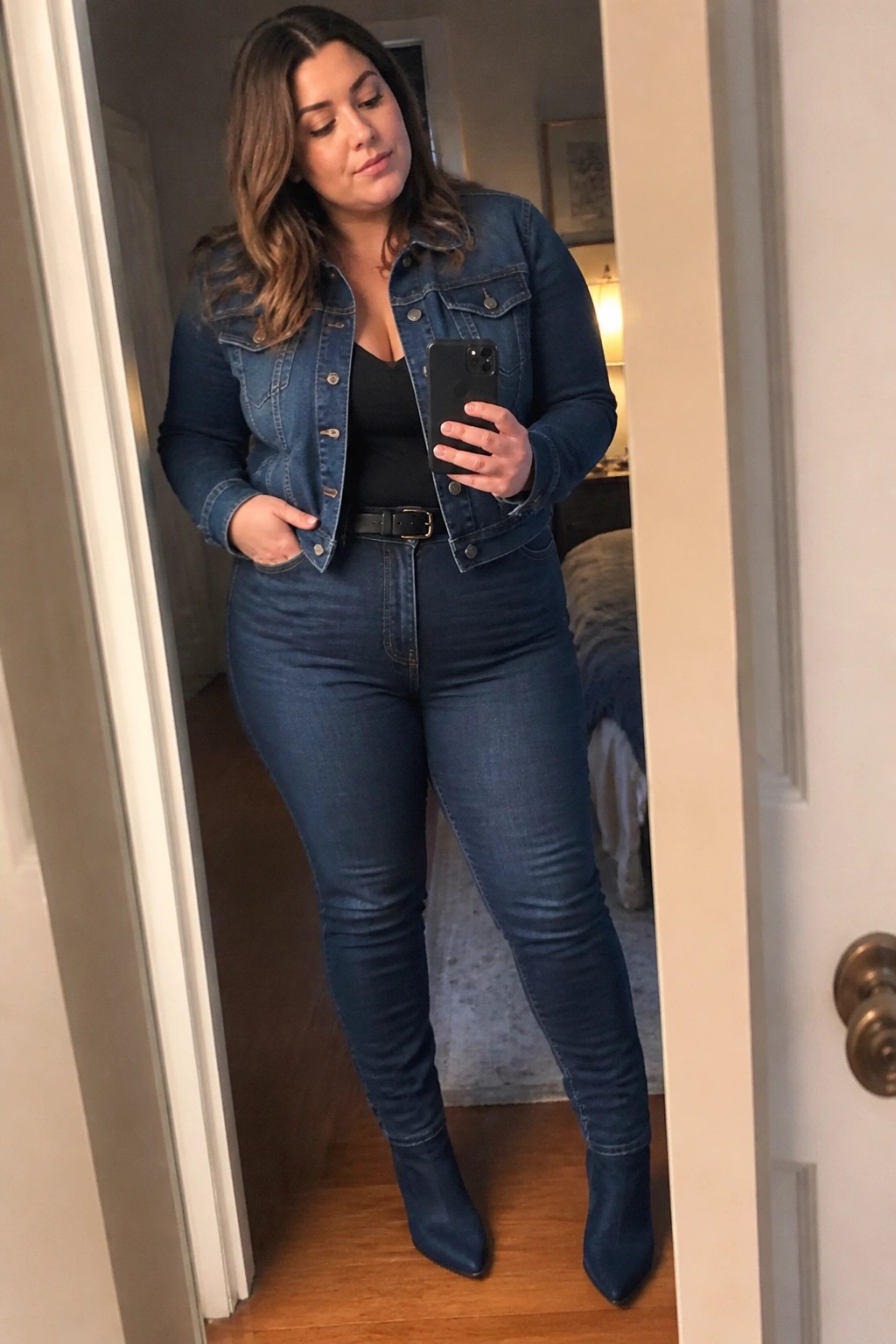 Woman wearing a cropped blue denim jacket over a black top, high-waisted blue jeans with a thin black belt, and matching blue pointed ankle boots while holding a phone in a mirror selfie.