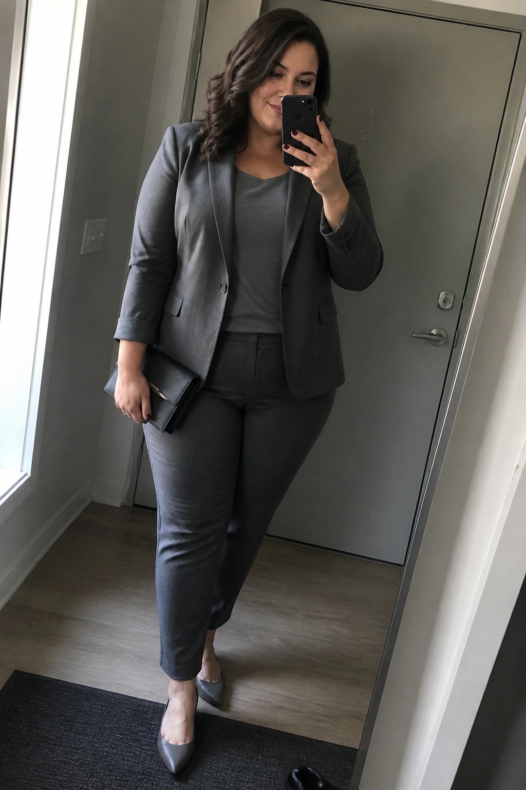 Woman wearing a full gray monochrome pantsuit with matching blazer, top, trousers, pointy flats, and clutch while taking a mirror selfie.