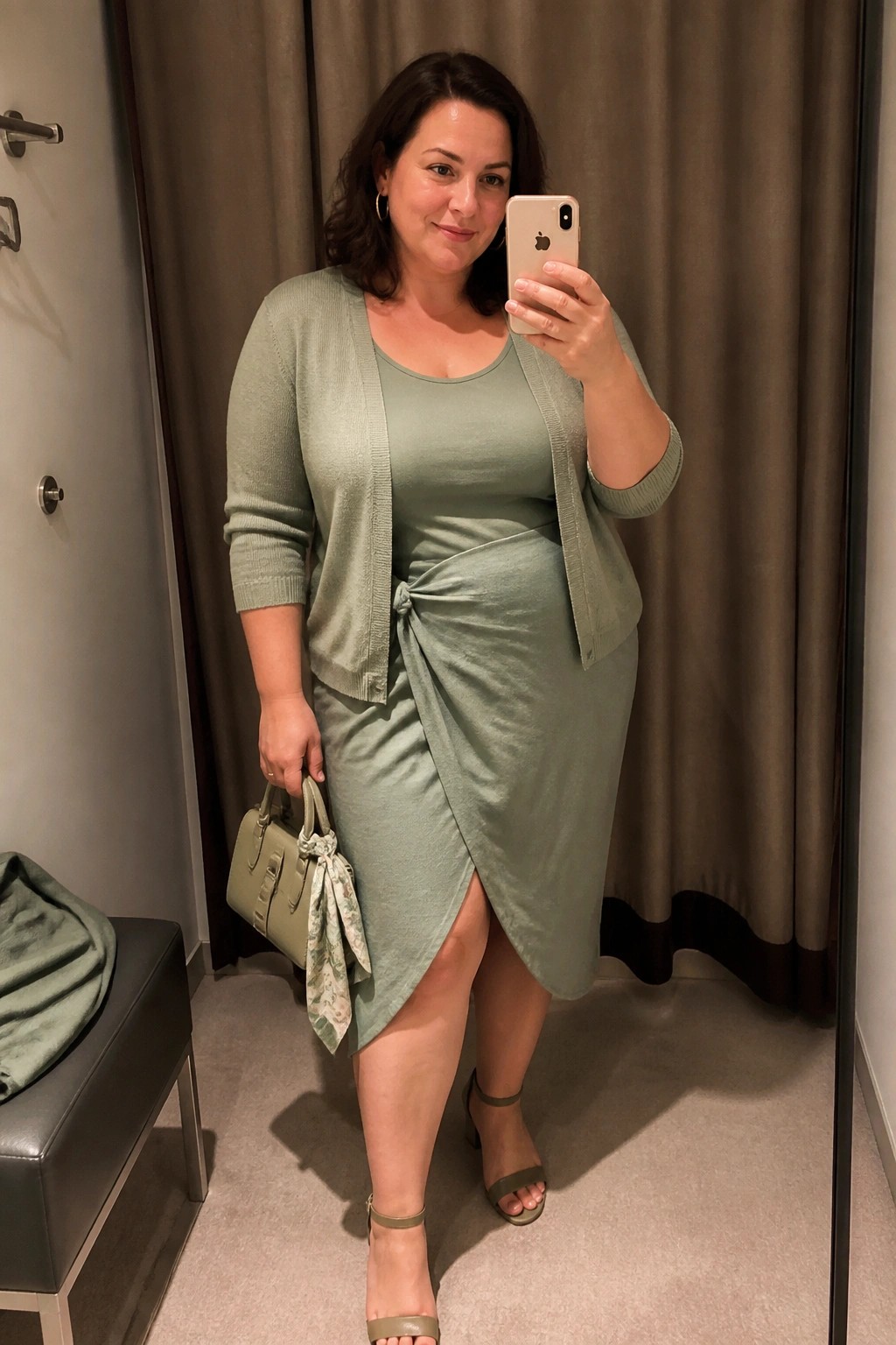 A plus size woman in a sage green monochromatic outfit featuring a sleeveless knit dress with side knot detail layered under an open matching cardigan paired with green block heel sandals and a coordinating handbag while holding a phone for a mirror selfie.