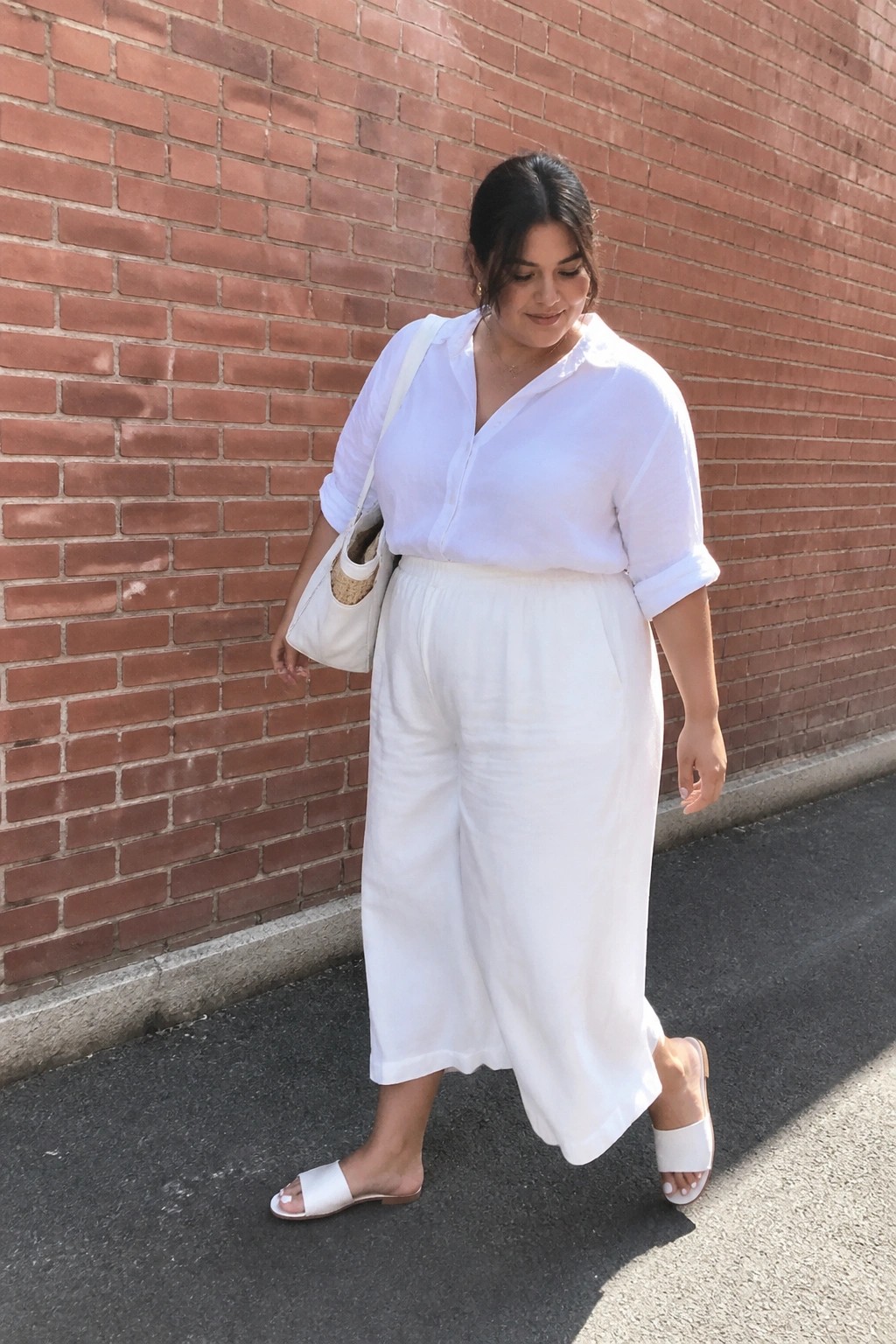 Woman in white shirt and pants carrying bag by red brick wall