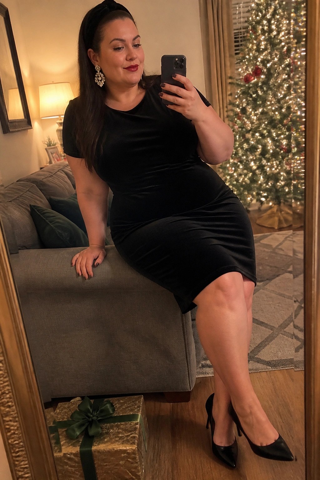 Woman in black velvet dress taking mirror selfie beside Christmas tree and gift
