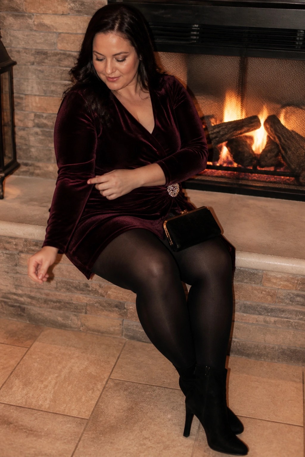 Woman in burgundy velvet dress with brooch seated by lit fireplace