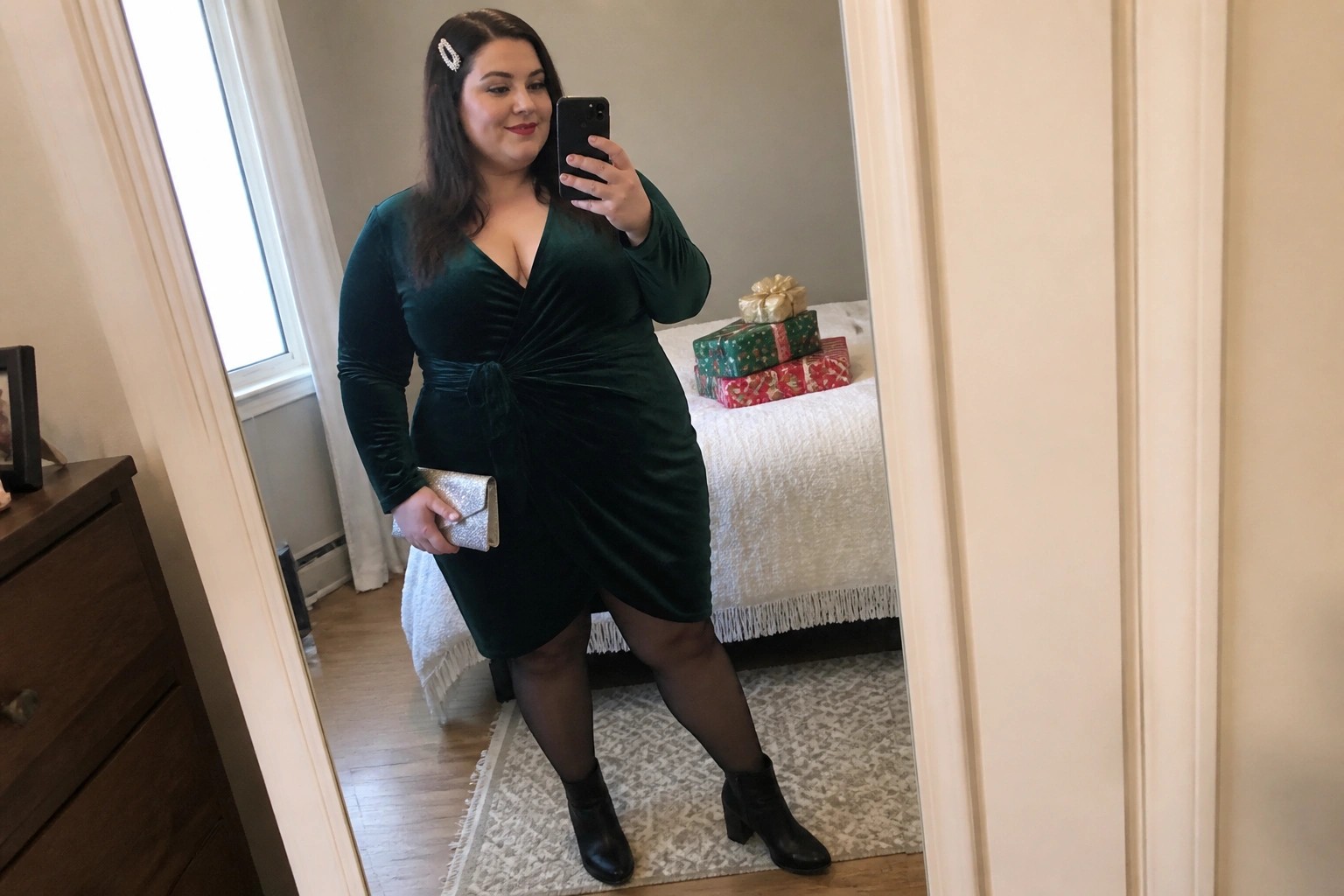 Woman in emerald green velvet dress taking mirror selfie in bedroom with gifts