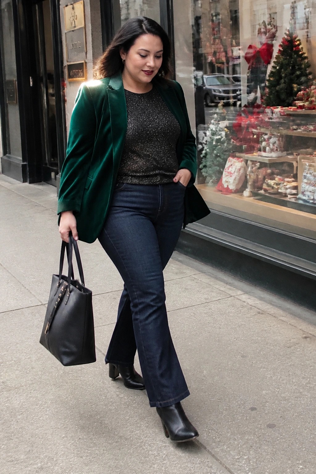 Woman in green velvet blazer and flared jeans holding black handbag on sidewalk