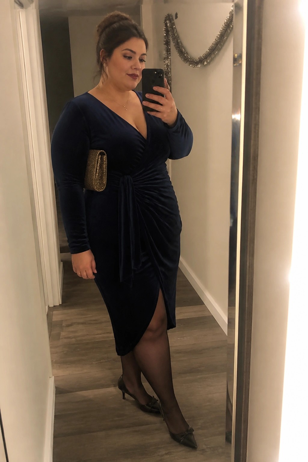Woman in navy blue velvet dress taking mirror selfie with gold clutch.