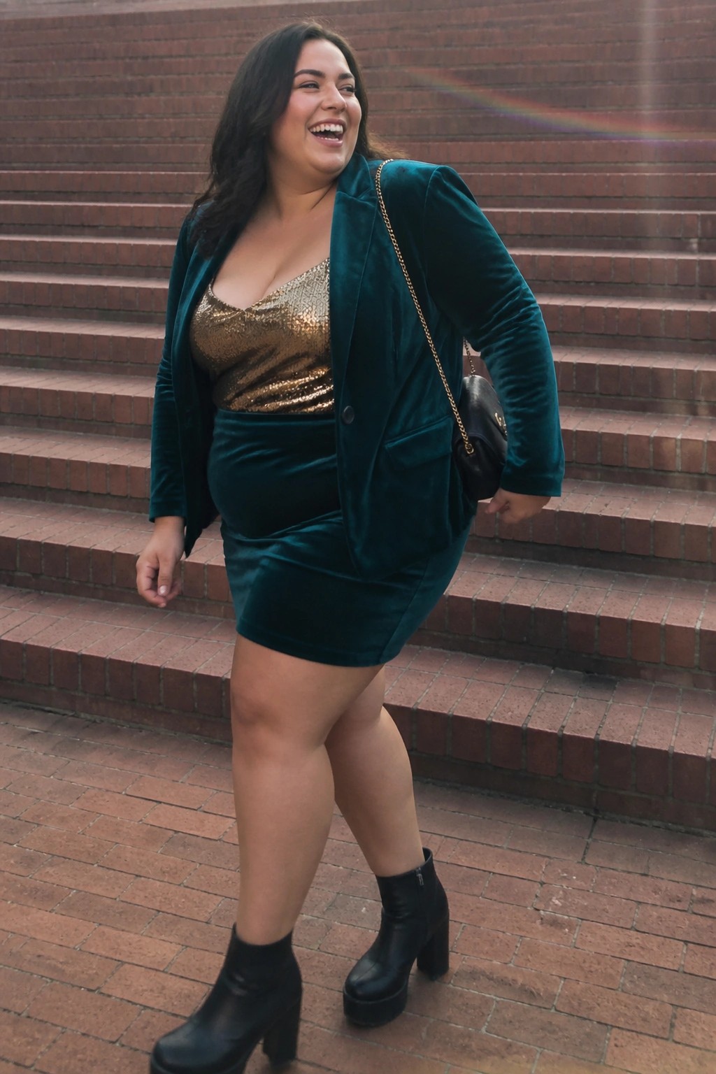 Plus size woman wearing teal velvet blazer and mini skirt over gold sequin camisole with black platform ankle boots and chain strap handbag.