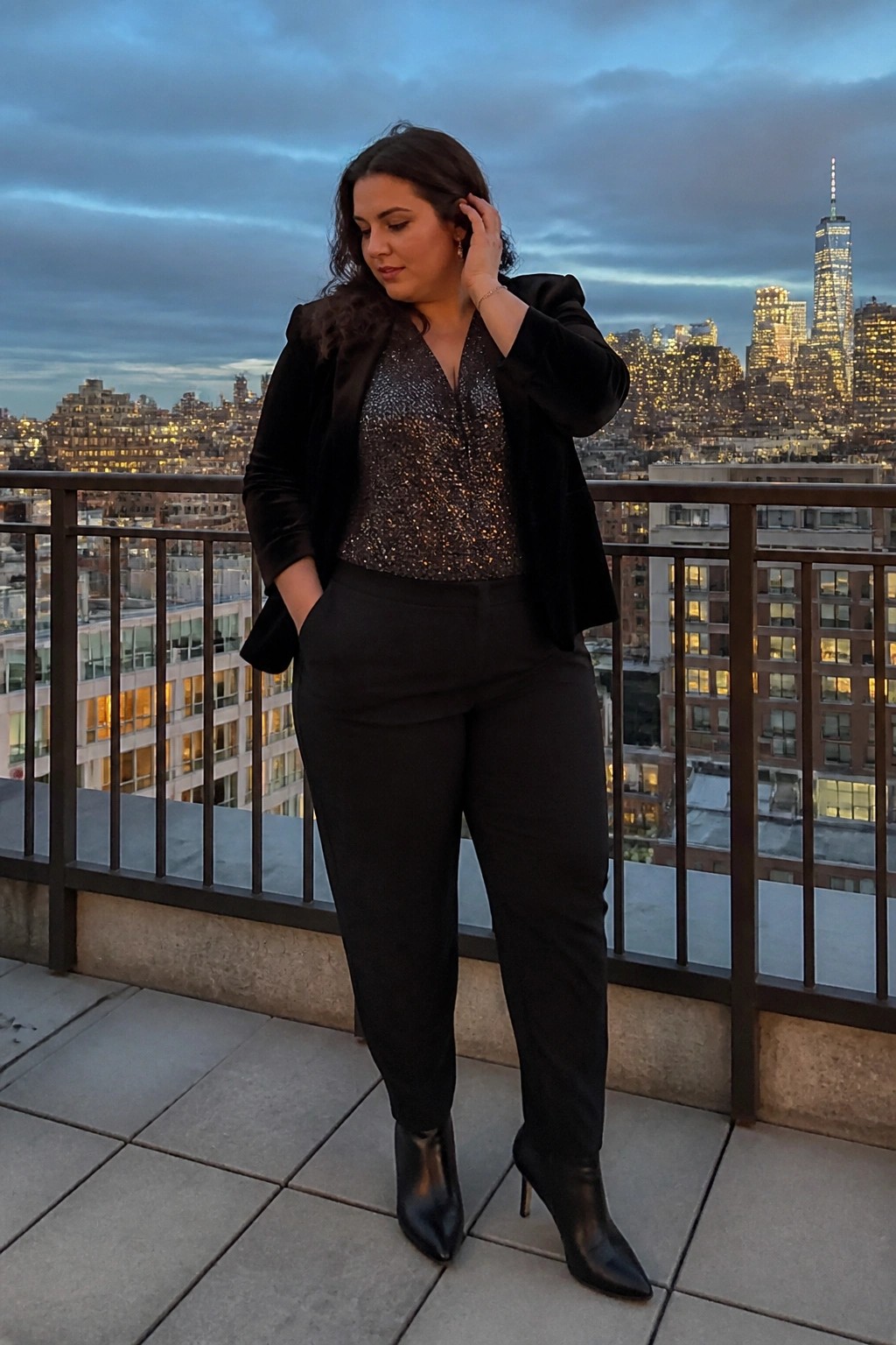 Plus size woman in black velvet blazer over sparkly sequin top paired with black trousers and pointed toe boots.