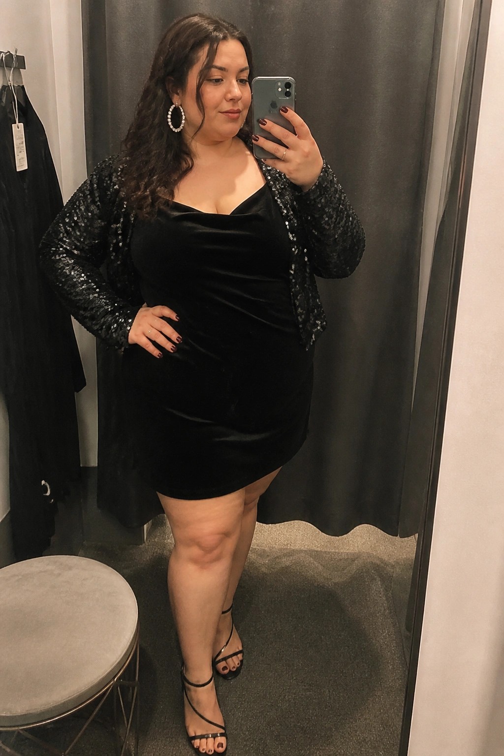 Curvy woman in black velvet dress and sequin jacket taking mirror selfie in fitting room.