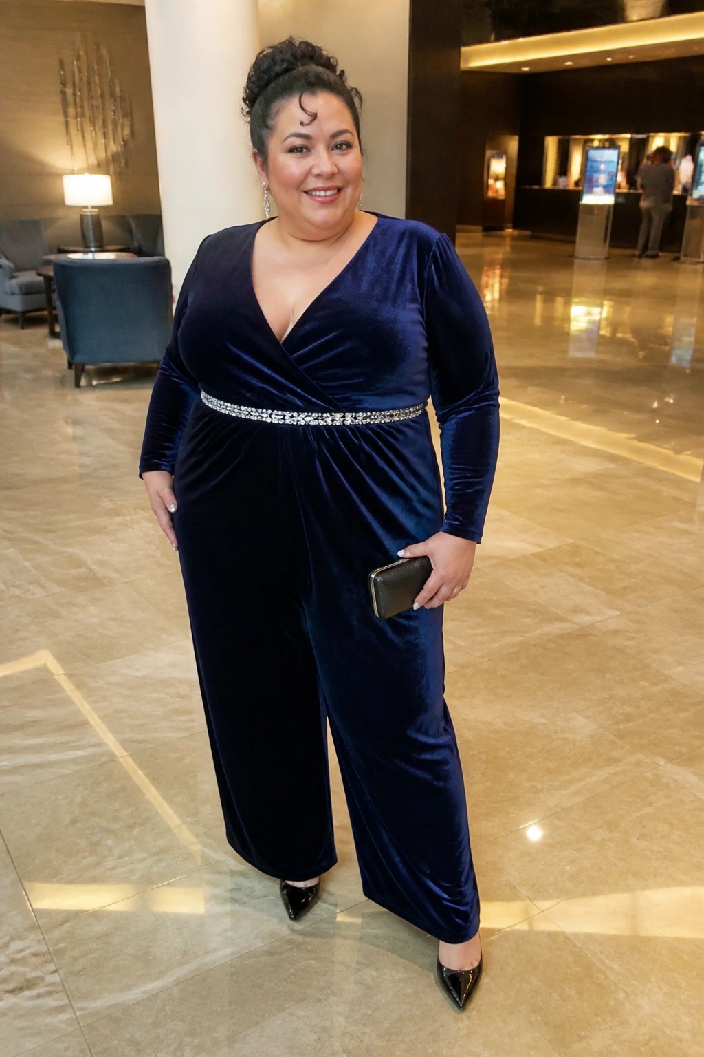 Plus size woman wearing a navy blue velvet long sleeve jumpsuit with a wrap neckline and rhinestone belt, holding a black clutch and wearing black patent heels in a hotel lobby.