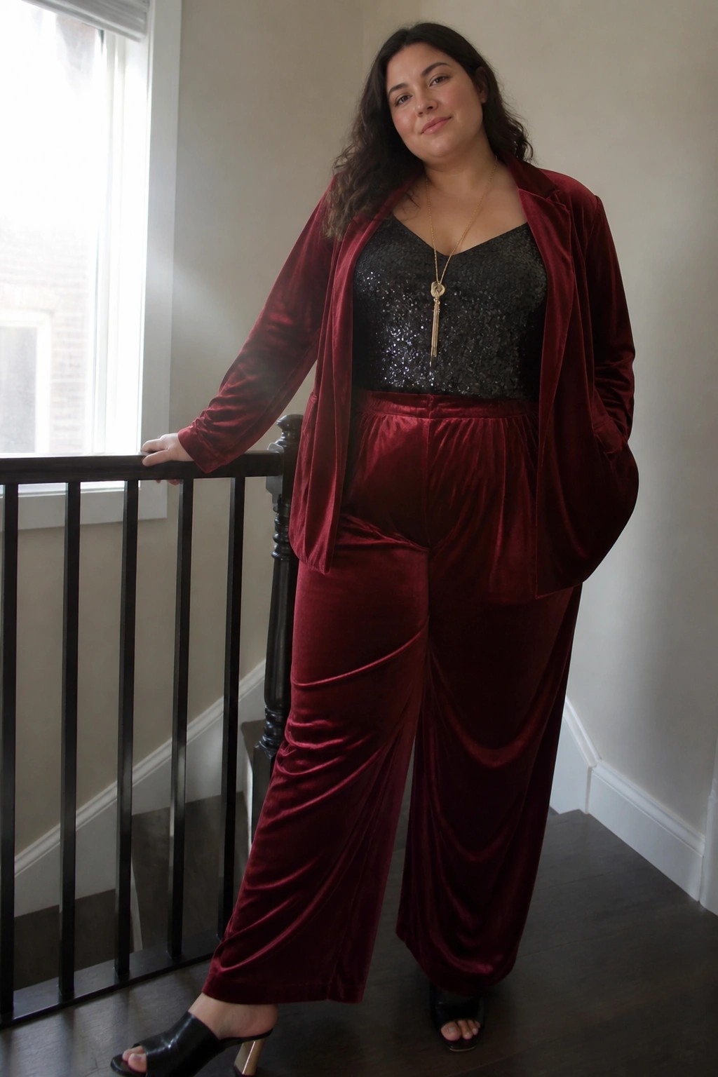 Plus-size woman in a burgundy velvet blazer and wide-leg pants paired with a black sequin camisole, gold pendant necklace, and black heeled mules.