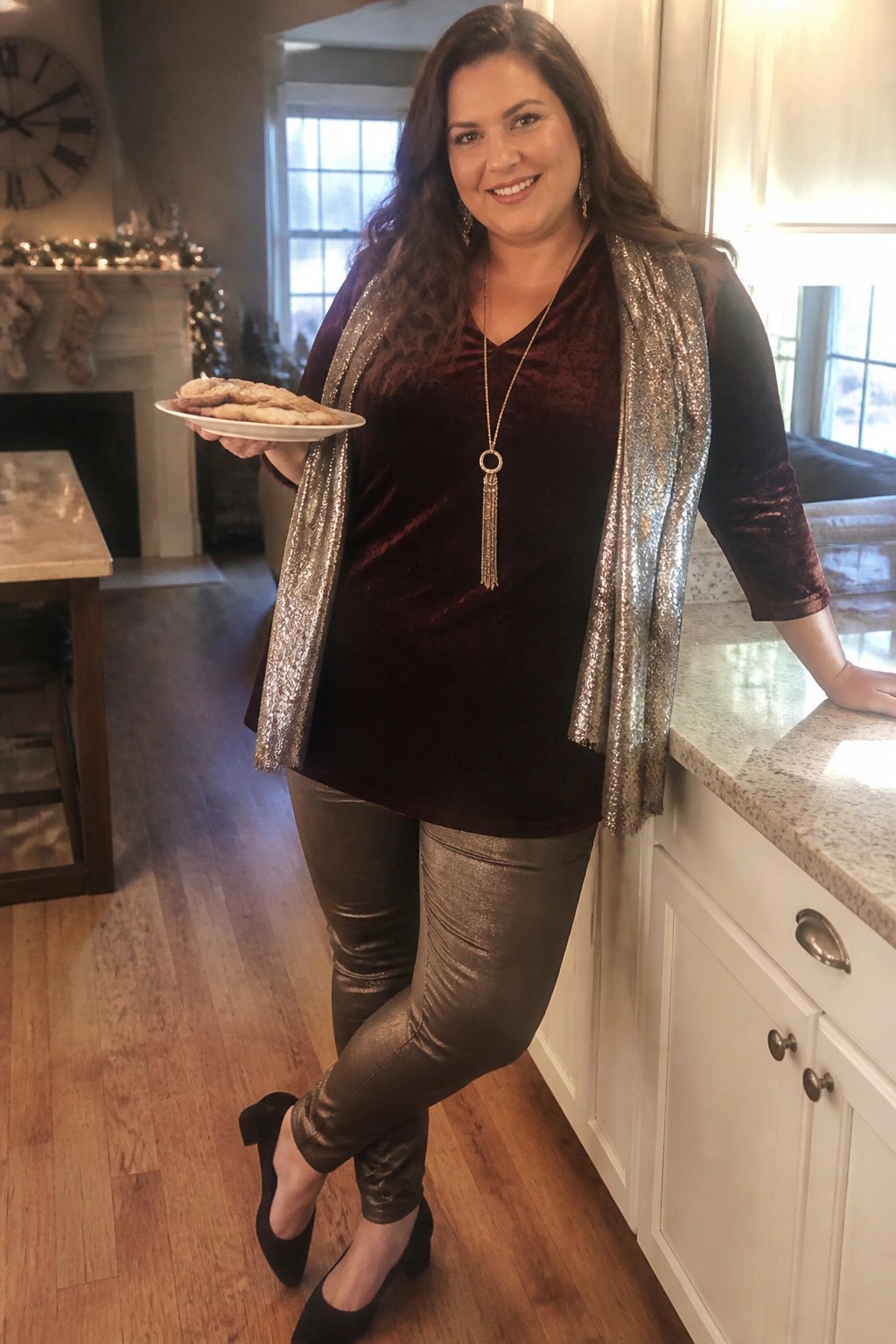 Woman in maroon velvet top and silver sequin vest holding cookies in kitchen.