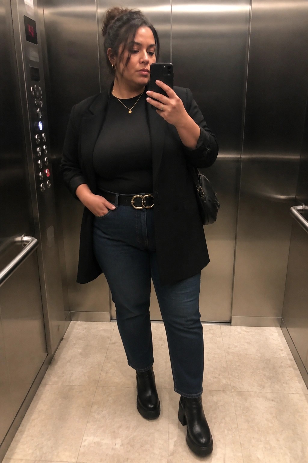 Woman in black blazer and jeans taking selfie inside metallic elevator.