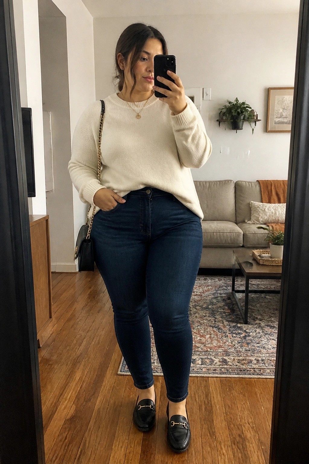 Woman in cream sweater and blue jeans taking mirror selfie in living room