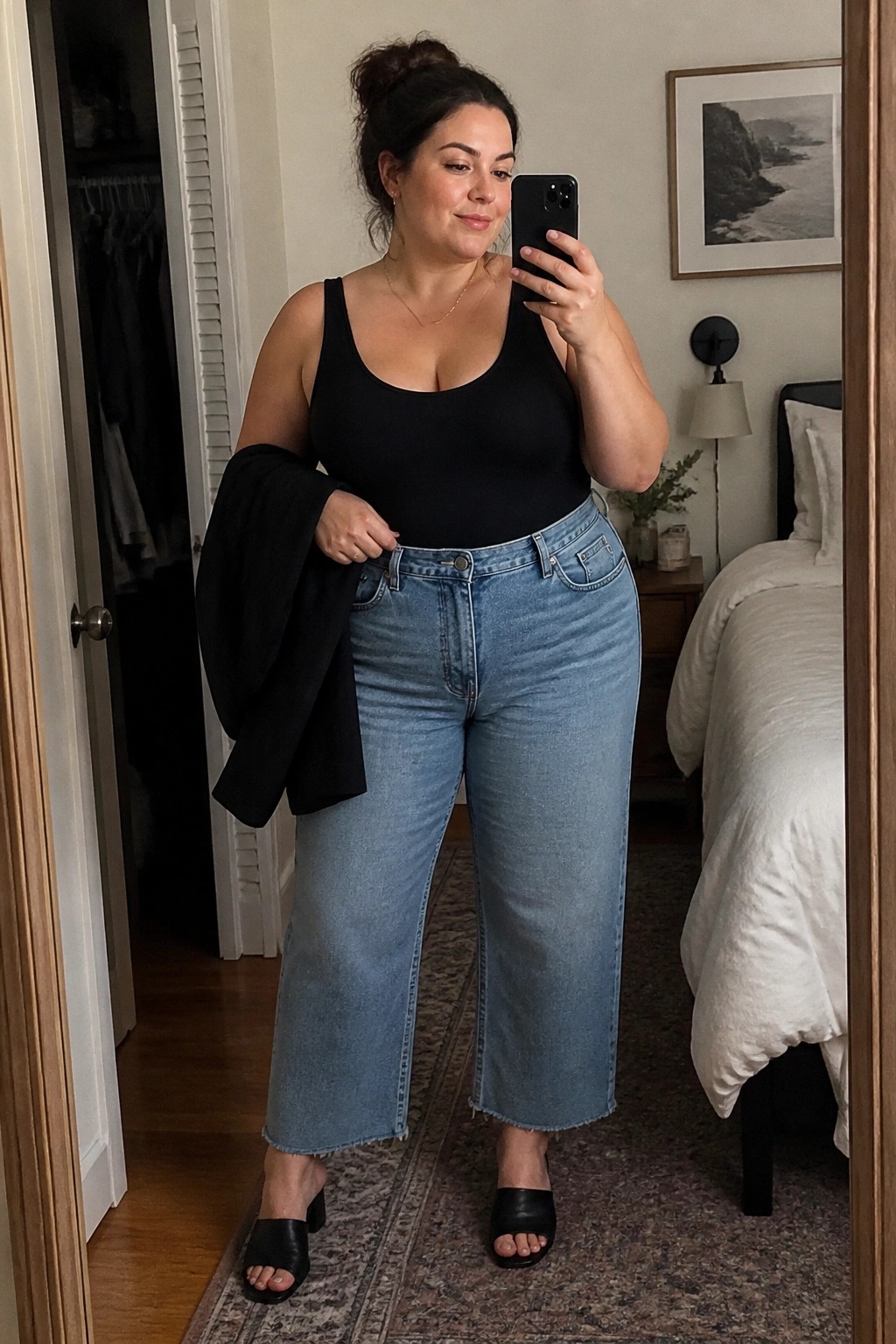 Woman in black tank top and light blue wide-leg jeans taking mirror selfie in bedroom
