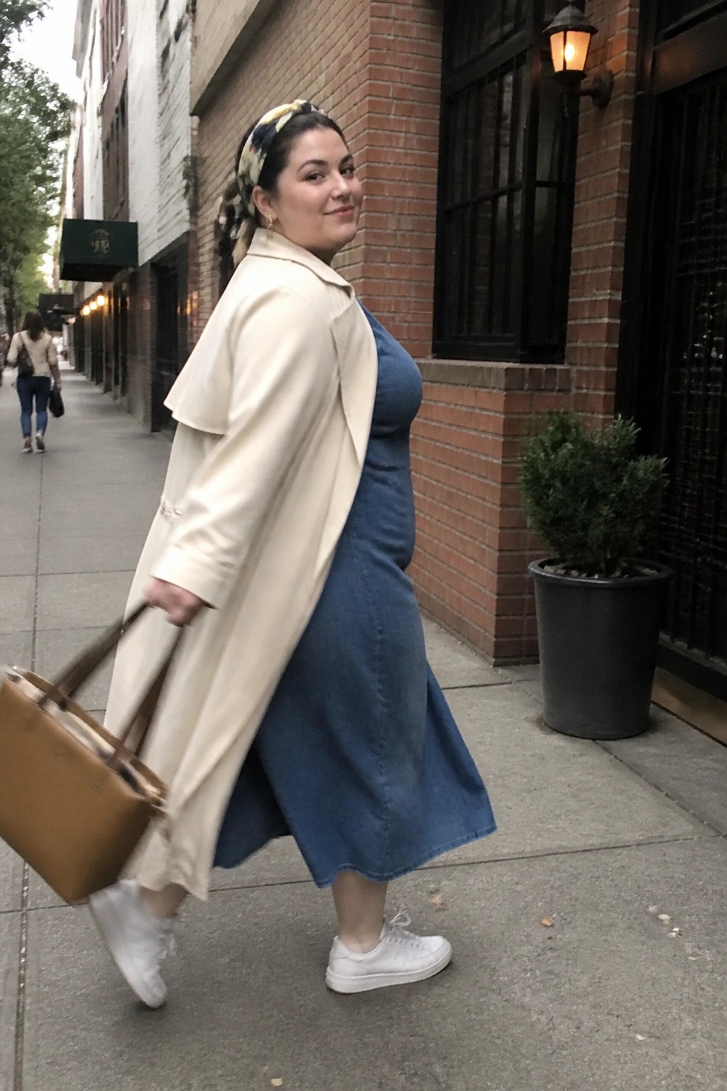 Woman in blue denim dress and beige coat carrying tote bag on city sidewalk