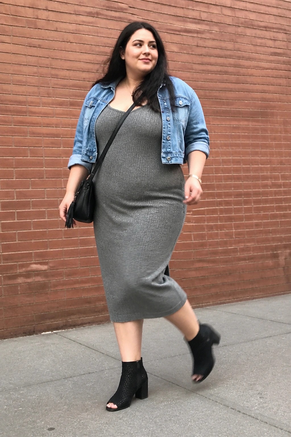 Woman in gray ribbed midi dress, denim jacket, and black boots by brick wall