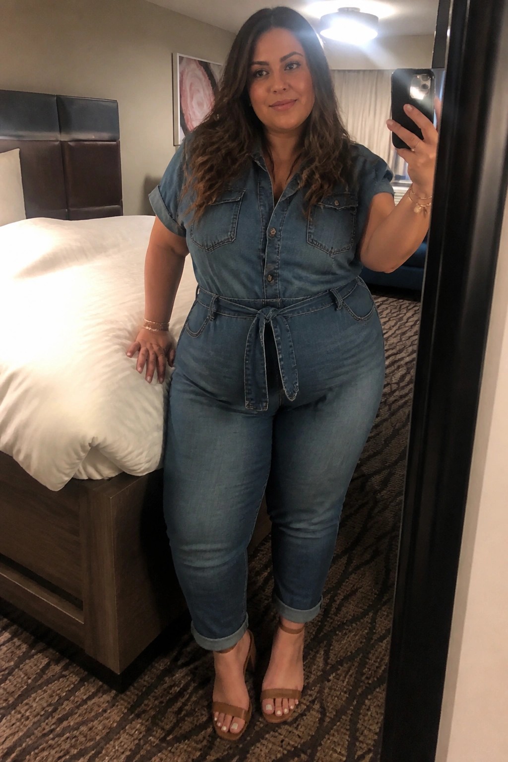 Woman in blue denim jumpsuit taking mirror selfie in hotel room beside bed
