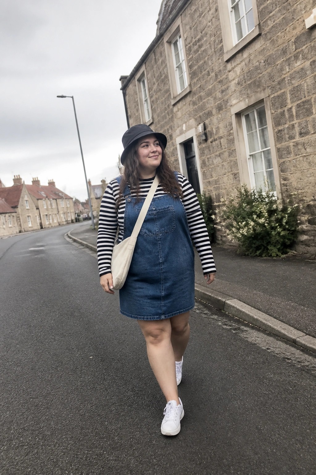plus size woman in denim overall mini dress over black white striped long sleeve top with white sneakers black bucket hat and beige crossbody bag