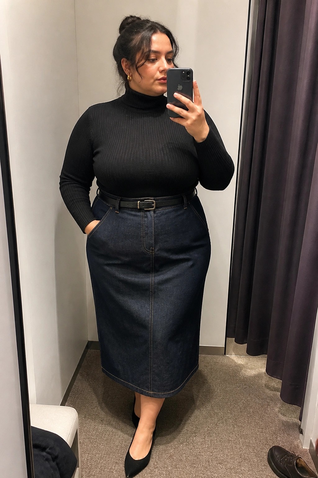 Woman in black turtleneck and long denim skirt taking mirror selfie in fitting room