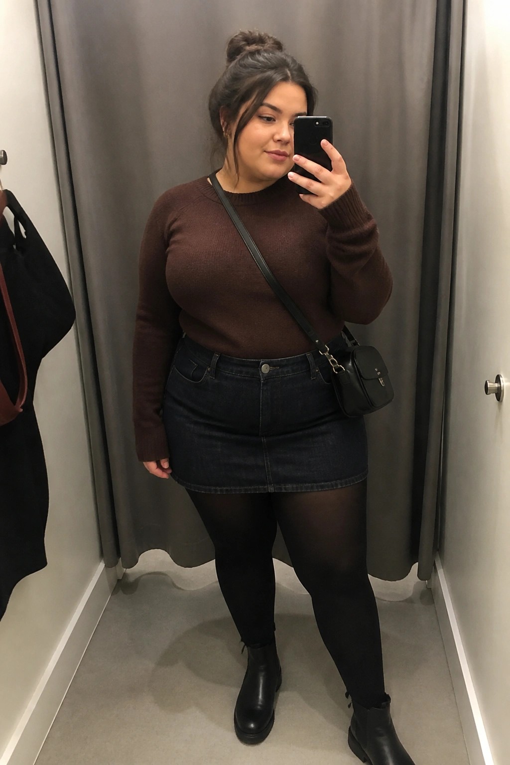 A plus size woman taking a mirror selfie in a fitting room, wearing a brown knit sweater tucked into a dark denim mini skirt, black tights, black ankle boots, and a small black crossbody bag.