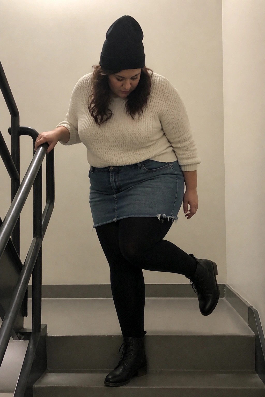 Woman in black beanie, cream sweater, and denim skirt on stairs holding railing.