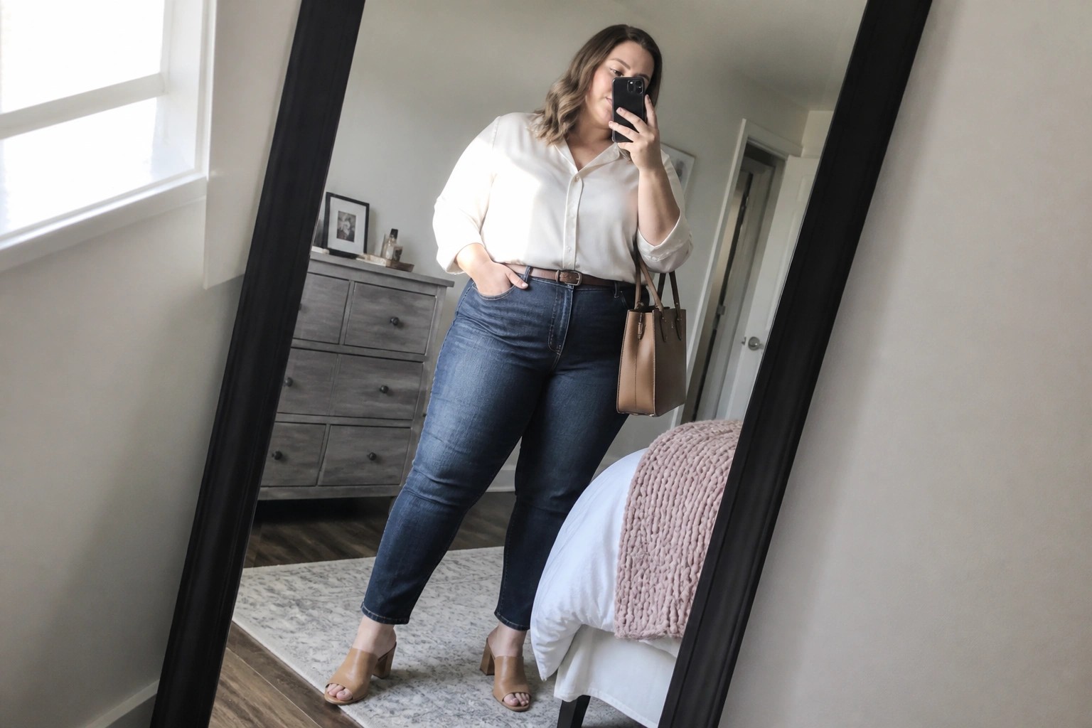 Woman in white shirt and blue jeans taking mirror selfie in bedroom