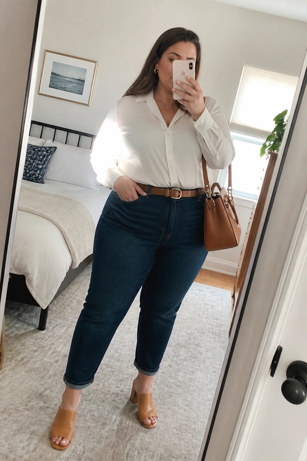 Woman in white shirt and blue jeans taking mirror selfie in bedroom
