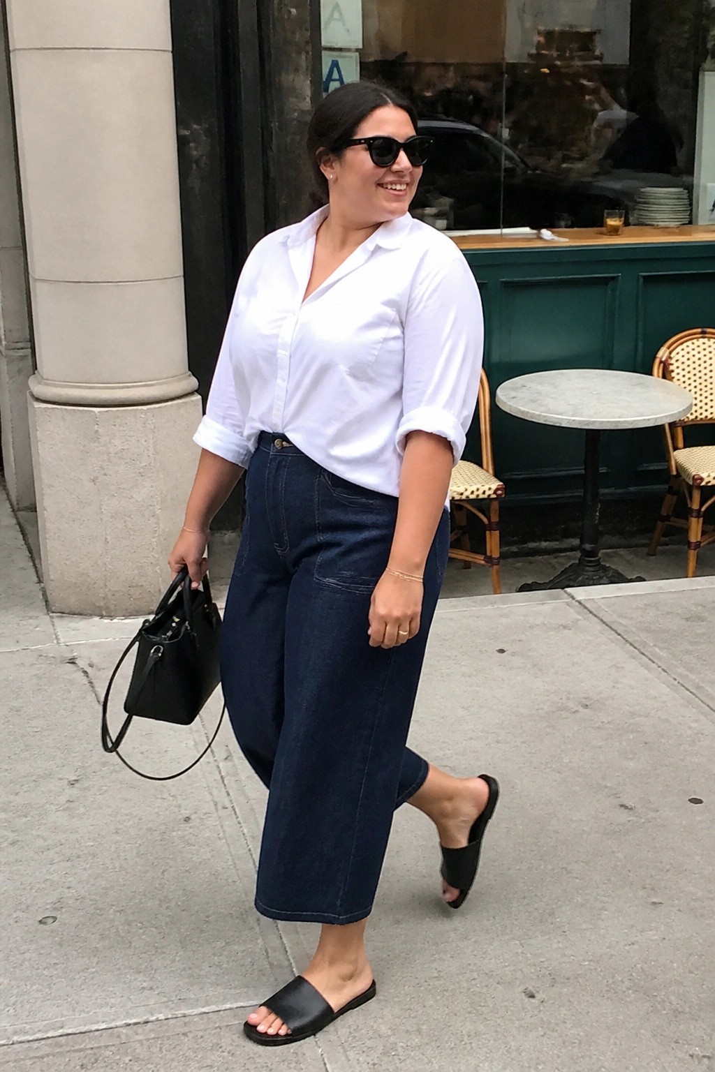 Plus size woman wearing a white button up shirt tucked into dark blue wide leg cropped denim jeans with black slide sandals and a black handbag.