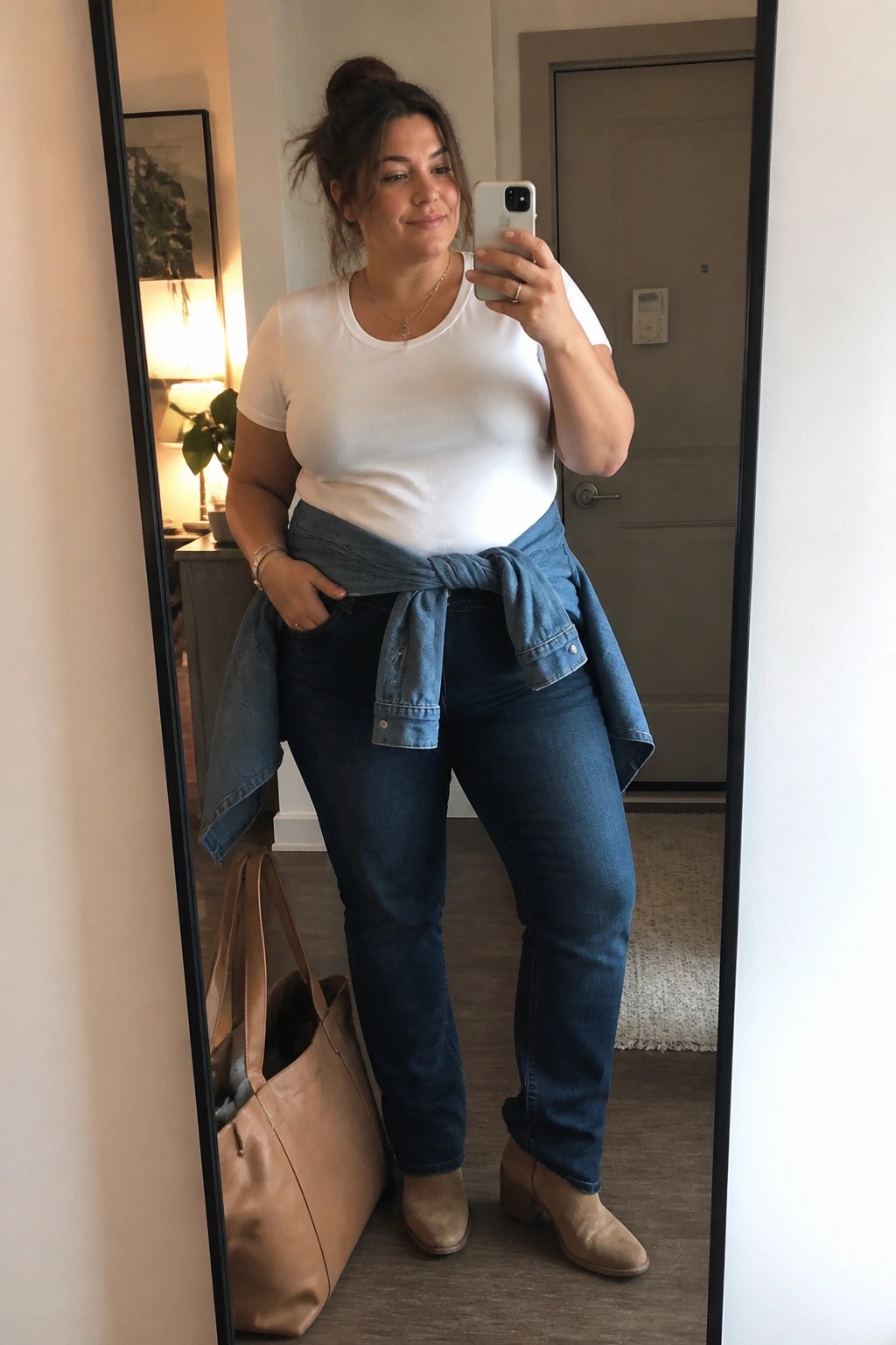 Plus size woman taking a mirror selfie in a white crewneck tee tucked into dark blue jeans with a denim jacket knotted at the waist beige ankle boots and a large tan leather tote bag visible in the reflection.