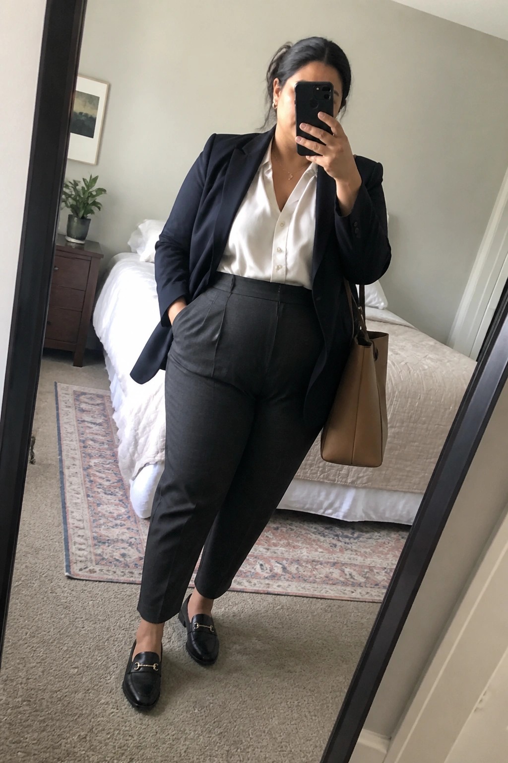Woman in black blazer, white shirt, and gray pants taking bedroom mirror selfie