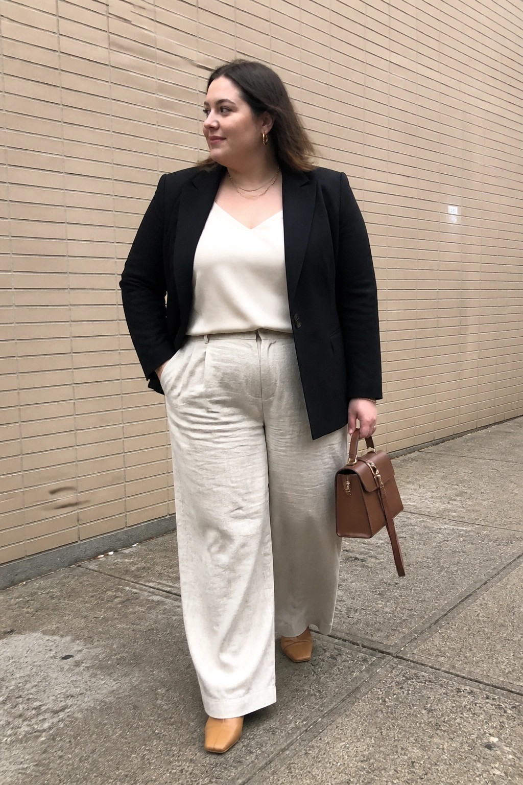 Woman in black blazer and beige wide-leg pants holds brown handbag by brick wall.