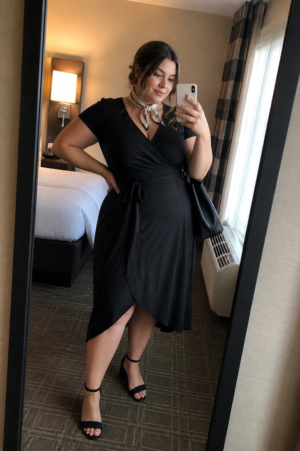 Plus size woman in a black short sleeve wrap dress with patterned neck scarf, black handbag, and ankle strap heels taking a mirror selfie.